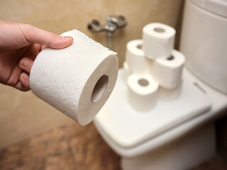 Man holding a roll of toilet paper against the background of a toilet with a lot of toilet paper on the lid.