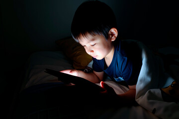 Asian boy child lying in bed using digital tablet, face illuminated by screen, holding device with both hands, focusing intently on display, partially covered by blanket