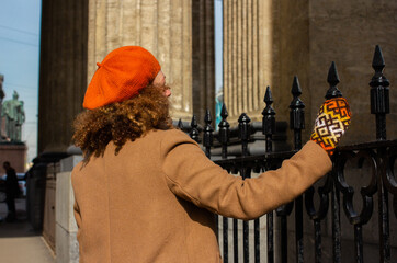 Stylish woman with curly hair in orange beret and beige coat stands by black wrought iron fence,...