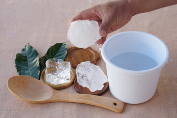 Hand holding natural alum crystal with a white plastic bucket of water. Concept of traditional water purification, natural minerals, and eco-friendly household cleaning using alum stones.