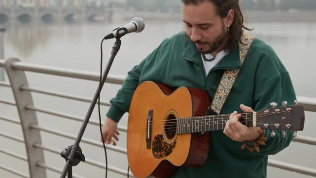 Medium shot of Caucasian male busker with dreadlocks, in green sweatshirt, playing song on acoustic guitar and singing into microphone, while performing live music for passersby on bridge