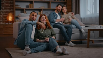 Naklejka premium Two diverse married couple four multiracial friends watching television at home party drinking beer alcohol cider drink in living room multiethnic multiracial men women focused enjoying movie film tv