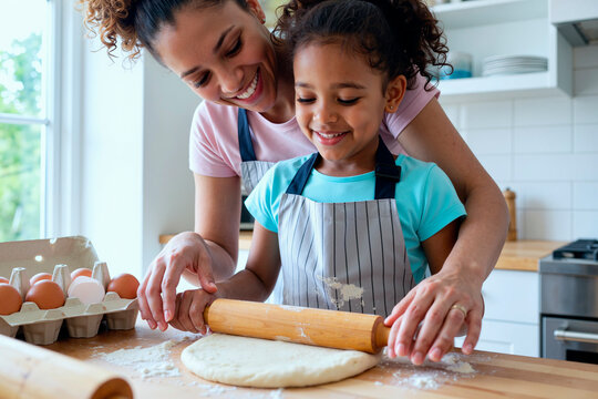 Cheerful woman helping young Black girl rolling dough with wooden rolling pin in kitchen, both smiling and enjoying baking activity together, eggs and flour visible on countertop