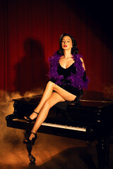 Performing artist in a purple feather boa poses on a grand piano with red curtains in the background during a glamorous evening event