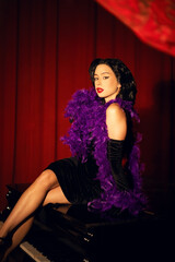 Performing artist in a purple feather boa poses on a grand piano with red curtains in the background during a glamorous evening event