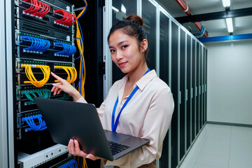 Young adult Asian woman standing in server room holding laptop, managing network cables and looking at camera, demonstrating IT professional working with data center equipment