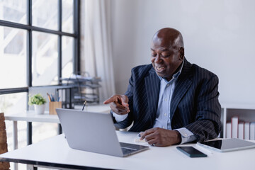Obraz premium Senior Black businessman having a video call on his laptop, actively discussing and pointing at the screen while working from his modern office in a pinstriped suit