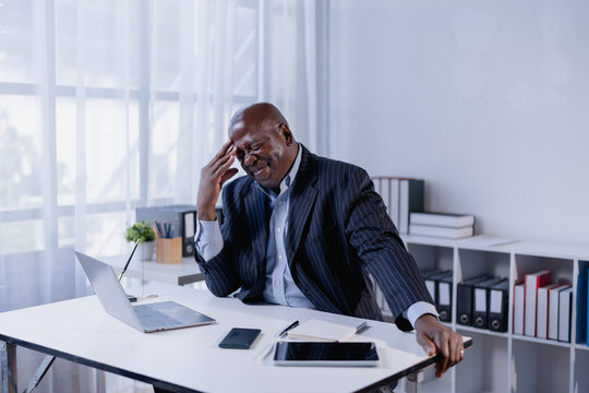African American businessman touching his temple, showing frustration and stress from work while sitting at his office desk with a laptop and other devices