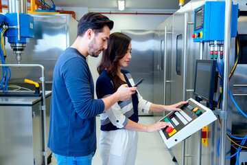 Young adult Caucasian man and young adult Asian woman operating industrial control panel in modern manufacturing facility, man holding smartphone while woman pressing buttons on machine