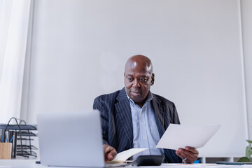 Black senior businessman wearing a pinstripe suit, sitting at an office desk with a laptop and calculator, carefully reviewing financial documents and a diary