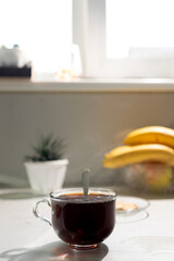 A clear glass cup filled with dark coffee rests on a kitchen counter. A spoon is in the cup. A plant, bananas, and a window are visible in the background.