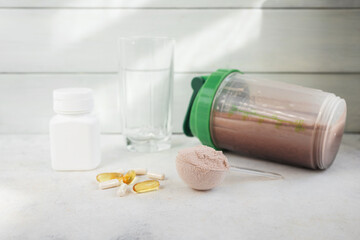 Chocolate whey protein powder, shaker with drink, omega-3 softgels, amino acid capsules, pill bottle and glass on light surface, fitness nutrition and supplement concept