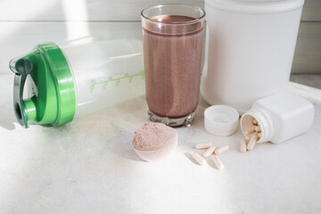 Scoop of whey protein powder, chocolate protein shake in glass, white capsules, shaker bottle and white plastic protein jar on light surface, fitness nutrition concept