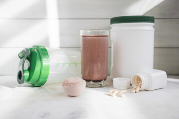 Scoop of whey protein powder, chocolate protein shake in glass, white capsules, shaker bottle and white plastic protein jar on light surface, fitness nutrition concept