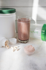 Scoop of whey protein powder, chocolate protein shake in glass, white capsules, shaker bottle and white plastic protein jar on light surface, fitness nutrition concept