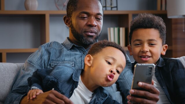 African American happy family with mobile phone smiling taking picture selfie grimacing father two kids sons boys children looking in smartphone trying funny photo mask recording video call at home