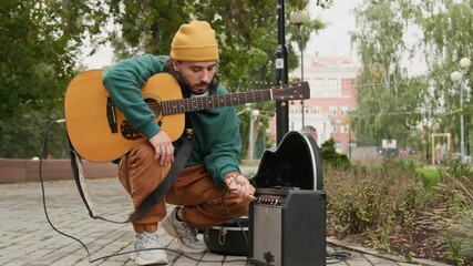 Full shot of Caucasian male busker in green sweatshirt and beanie crouching on sidewalk in public park with guitar, plugging electric cord into amplifier or audio speaker for live street performance