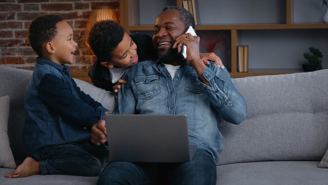 Happy African American family father working sons disturb dad talking mobile phone laptop using finish work laughing with two little boys children need attention kids noisy distracting daddy at home 