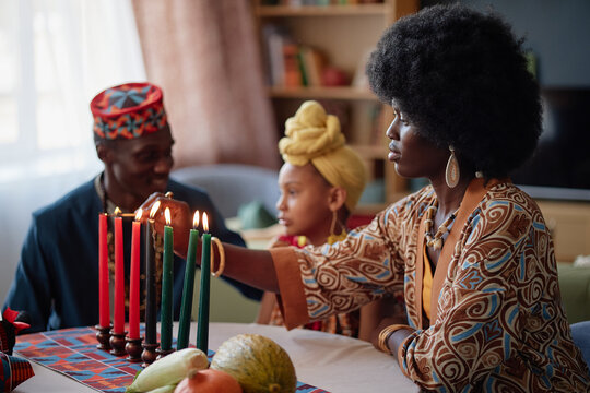 Black woman lighting kinara candles while sitting at table with Black man and Black child during Kwanzaa celebration, all wearing traditional accessories, fruits on table