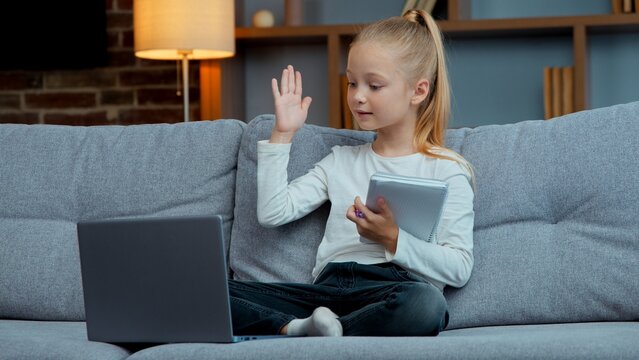 Caucasian kid child girl daughter e-learning class on sofa writing notes raising hand answer to teacher write study and looking laptop during virtual online lesson in cozy living room evening home