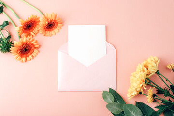 Envelope with blank card, orange gerbera flowers and yellow chrysanthemums on coral pink background. Floral mockup for message or invitation. Top view, flat lay