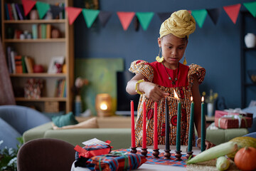 Black girl child lighting kinara candles during Kwanzaa celebration, standing at table with...