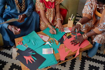 Black woman, Black man, and Black child creating colorful paper hand cutouts together at table, celebrating Kwanzaa with arts and crafts, hands actively drawing and cutting shapes