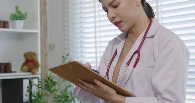 Asian female pediatrician looks down while writing on clipboard in clinical office during early care session showing focus attention to detail and professionalism in medical documentation process