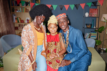 Black woman, Black man, and Black child sitting together smiling, wearing traditional clothing and jewelry, celebrating Kwanzaa with festive decorations in background