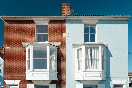 Semi detached British houses one with a pastel blue painted brick wall, the other unpainted. Located at a seaside town, the large Georgian bay windows enjoy sea views.