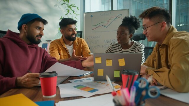 Focused concentrated informal casual startup business team considering paper document report information together talking solving finance problem multiethnic four people in office meeting with laptop