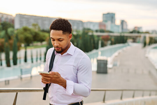 Portrait of a young businessman man using a smartphone mobile phone walking down the street, surrounded by moder corporate office buildings and modern city architecture