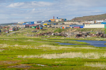 Riverbank view in Anadyr, Chukotka, Russia, with water, grassy tundra, and colorful village buildings under summer daylight.

