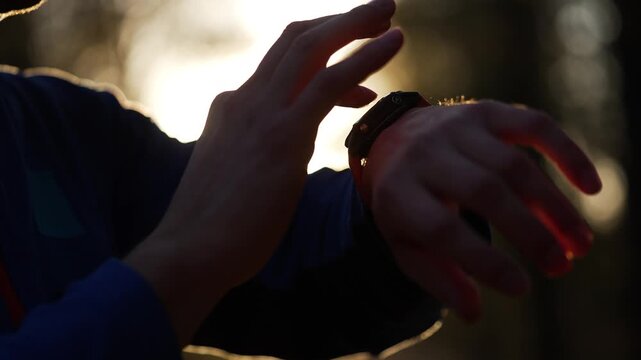 Silhouette of a runner checking workout results on a smartwatch, a close-up of his hands against the bright sun. The athlete is tracking his fitness progress after an outdoor training session
