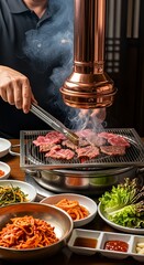 Close-up of hands grilling thin slices of Korean BBQ beef on a tabletop grill with a copper exhaust hood, surrounded by traditional side dishes (banchan).