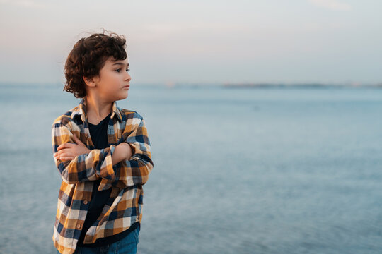 A contemplative child with crossed arms stands by the sea, gazing thoughtfully at the horizon as the sky turns pastel shades at dusk.