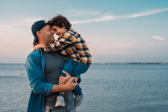 A joyful father and son embrace by the seaside, sharing smiles under a pastel sky.