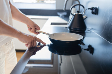 Close-up of man in white clothes putting frying pan on electric stove in modern kitchen. Concept of cooking, daily routine, and home lifestyle. Symbol of preparation, comfort, minimalistic interior