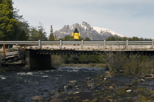 Person in Yellow Jacket Sitting on a Bridge Over a River with Snowy Mountain - Powered by Adobe