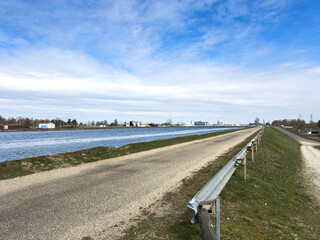 View of the Rhine River in France with a gravel path and metal guardrail alongside waterway under a partly cloudy blue sky with industrial buildings