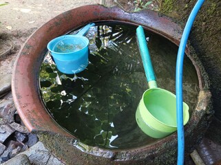 Clay water jar filled with clear water and two plastic dippers in blue and green. Natural and traditional outdoor setting with peaceful rural atmosphere.