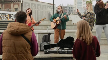 Full shot of two young Caucasian male and female street musicians playing guitars and singing popular songs for eager public, passersby listening, singing along, filming performance on smartphones