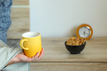 Relaxed morning scene — woman lying in bed holding a yellow cup of coffee. A wooden bedside table with an alarm clock and cookies adds cozy warmth to the room.