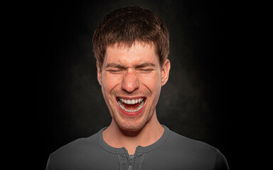 A young man with brown hair, wearing a gray shirt, screaming or laughing intensely with his eyes closed and mouth wide open against a dark background.