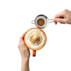A hand sprinkles cocoa powder over a frothy cappuccino in an orange mug. The background is transparent, highlighting the coffee preparation process.