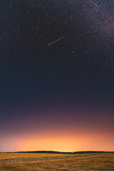 Natural Night Starry Sky Above Field Meadow. Glowing Stars, Meteorite Trail And Sunset Sunrise Lights Above Landscape In August Month. Meteoric Track In Night Sky Background