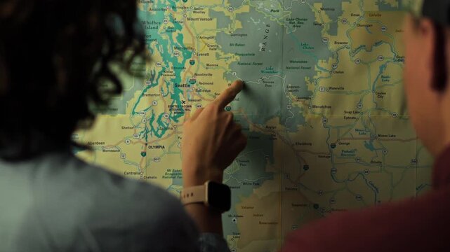 Individuals are focused on a map showing a forest and lake region, pointing to specific locations and discussing routes during a sunny afternoon, indicating an outdoor adventure plan