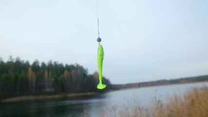 Close up bright green soft plastic fishing lure with hook, sinker spinning on fishing line against blurred background of autumn lake and forest, showing fishing tackle. Fishing lure spinning on line