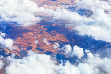 Fluffy clouds drift over sunlit, arid terrain, revealing reddish patterns that hint at ancient land formations. Aerial view captures striking contrast between sky softness and earth rugged texture