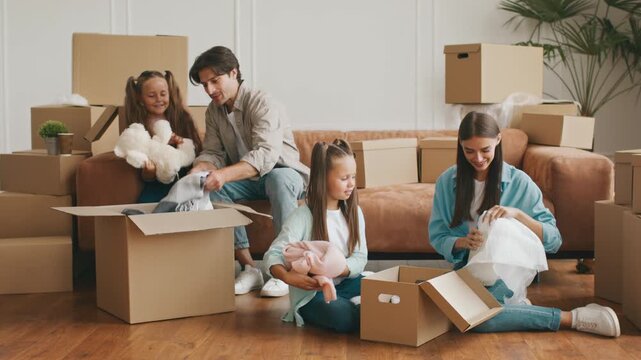 A Caucasian family is happily unpacking boxes in their new apartment. Two children help their parents organize clothes while smiling and enjoying the moment after moving.